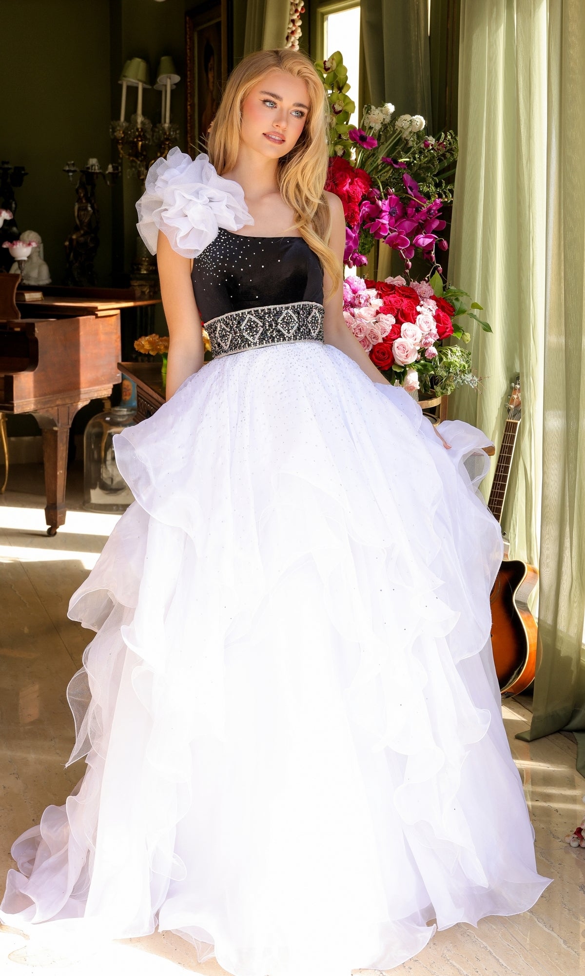 A young woman poses indoors wearing the Long Prom Dress 39557 by Ava Presley, a one-shoulder ball gown with a black bodice, white layered skirt, and floral shoulder detail. Flowers, a piano, and a guitar are visible in the background.